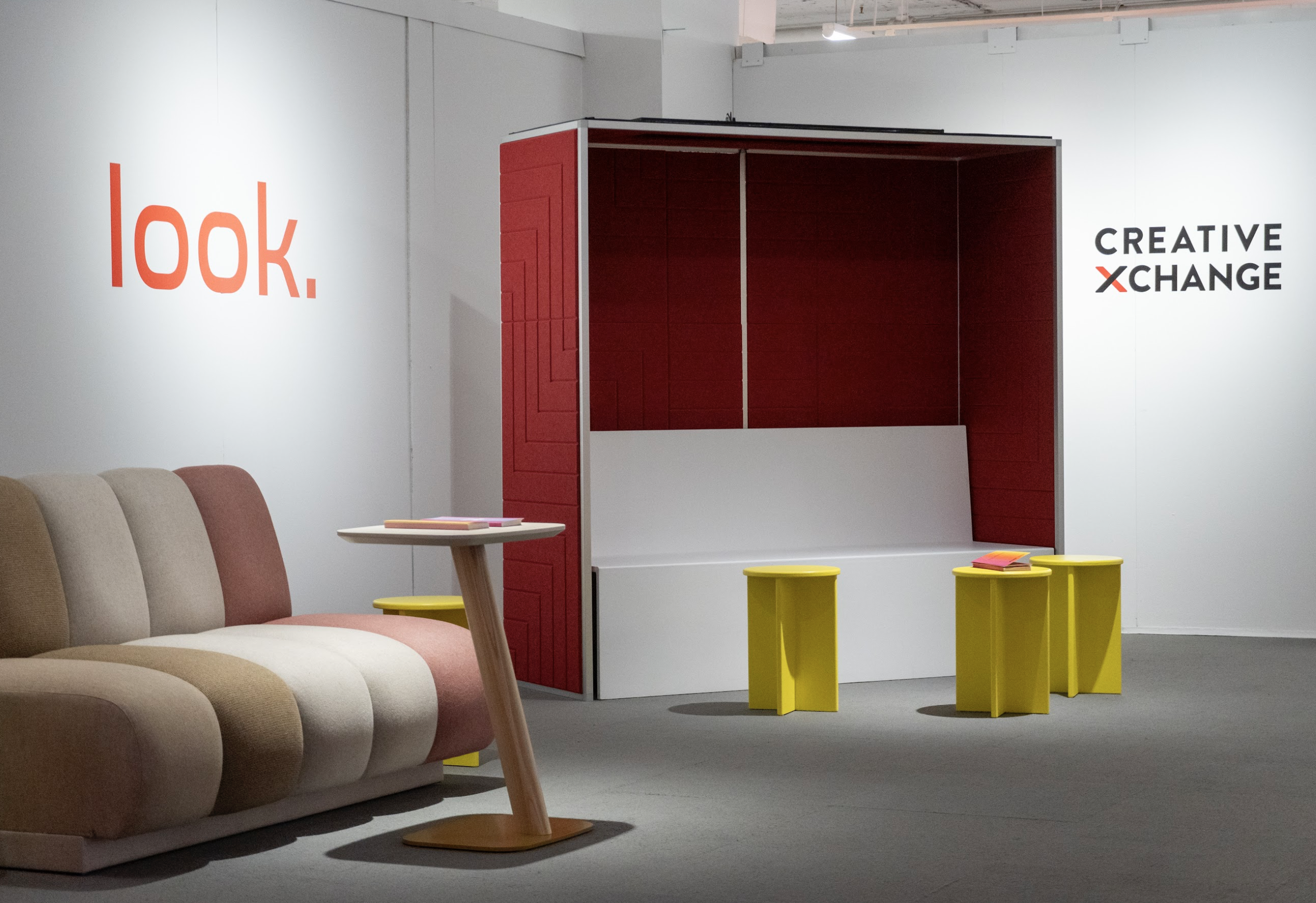 An exhibition space featuring a clean, modern seating arrangement with a beige, brown, and pink striped sofa to the left, a small round side table in front, and a booth-style red and white bench with three small bright yellow stools centered beneath bold signage reading 